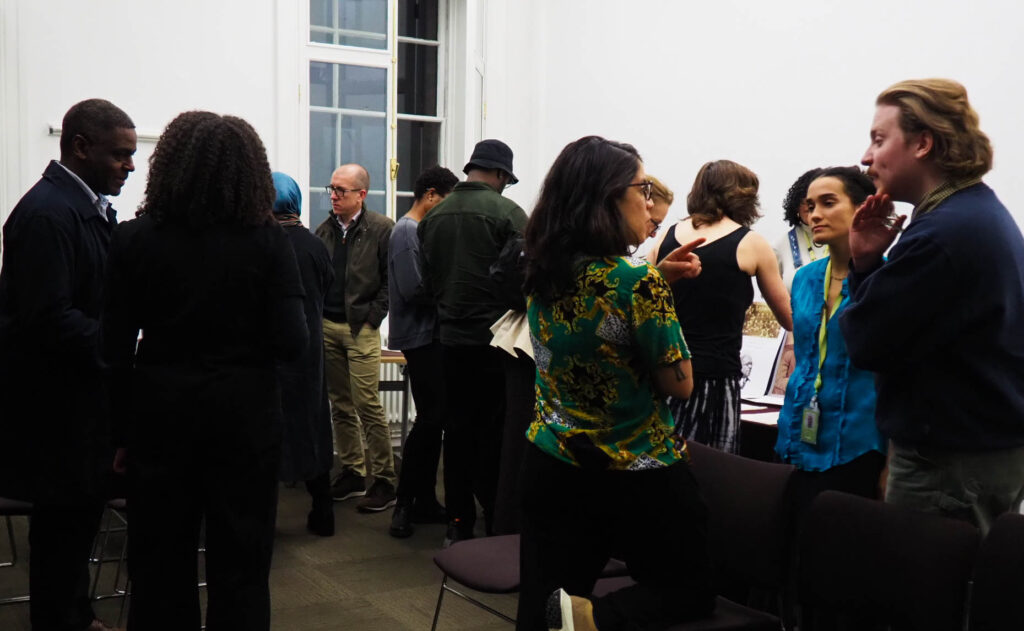 A crowd of people stand talking in separate groups inside a white room. Some rows of chairs are visible, but no one is sitting.