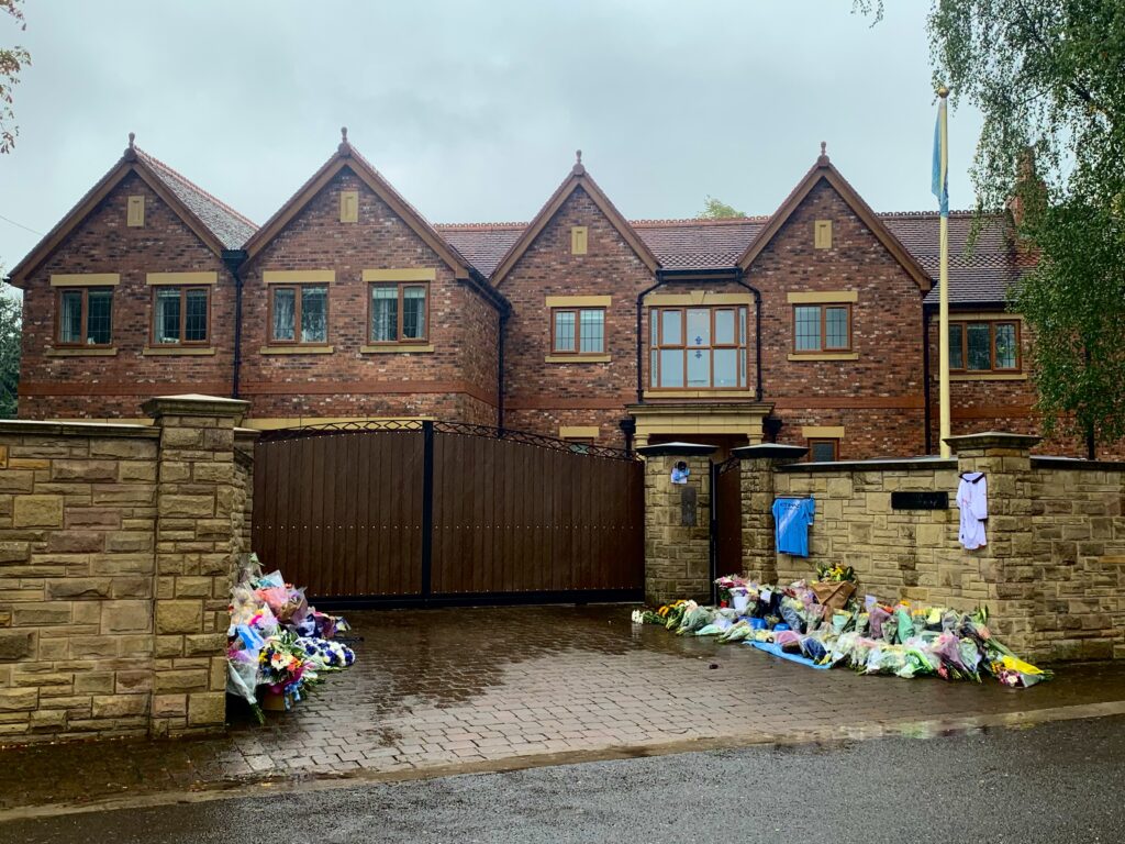 A large brown bricked mansion and a gated driveway. Flowers, wreaths, and blue Manchester City football shirts are strewn across either side of the driveway. Everything is wet from recent rain.