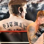 Boxer Ricky Hatton is a white man with a black tattoo sleeve and wearing black boxing gloves. He is standing sideways with his fists to his face and smiling. Behind him is a large poster of himself with his back turned. ‘HITMAN’ is tattooed on his back.