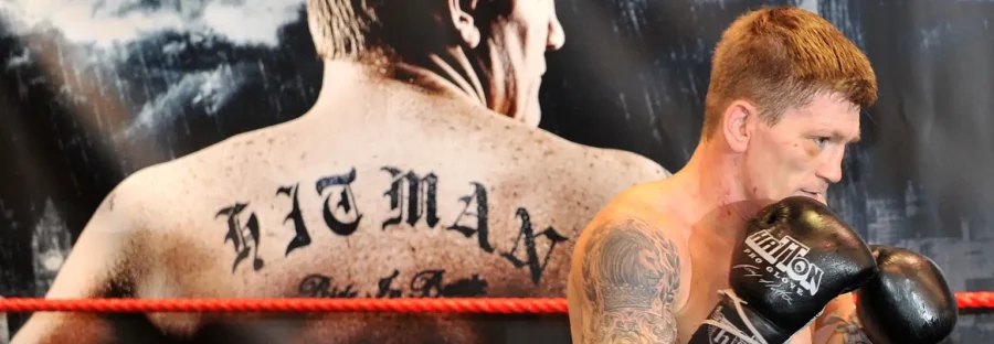 Boxer Ricky Hatton is a white man with a black tattoo sleeve and wearing black boxing gloves. He is standing sideways with his fists to his face and smiling. Behind him is a large poster of himself with his back turned. ‘HITMAN’ is tattooed on his back.