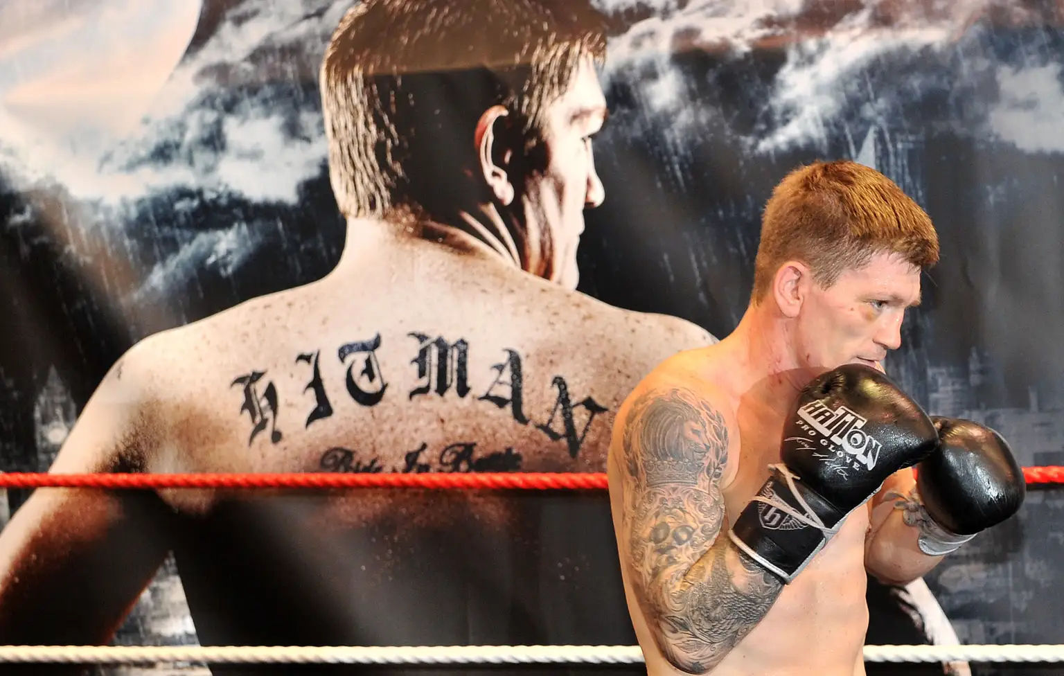 Boxer Ricky Hatton is a white man with a black tattoo sleeve and wearing black boxing gloves. He is standing sideways with his fists to his face and smiling. Behind him is a large poster of himself with his back turned. ‘HITMAN’ is tattooed on his back.
