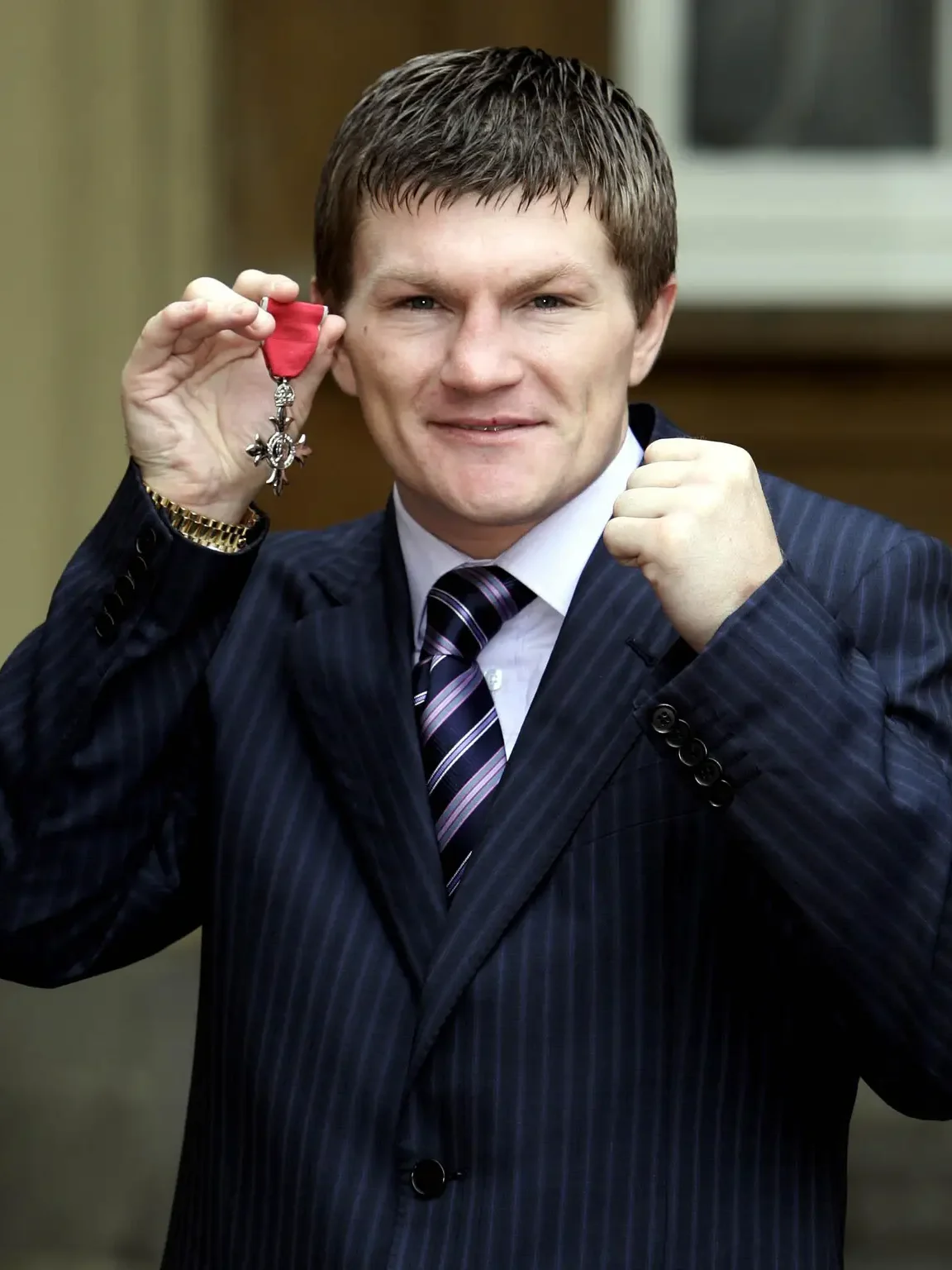 Ricky Hatton is a white man with short blond hair, wearing a black suit, white shirt, and blue-and-silver striped tie. He is smiling. In his hand he holds an MBE: a gold cross with red ribbon.