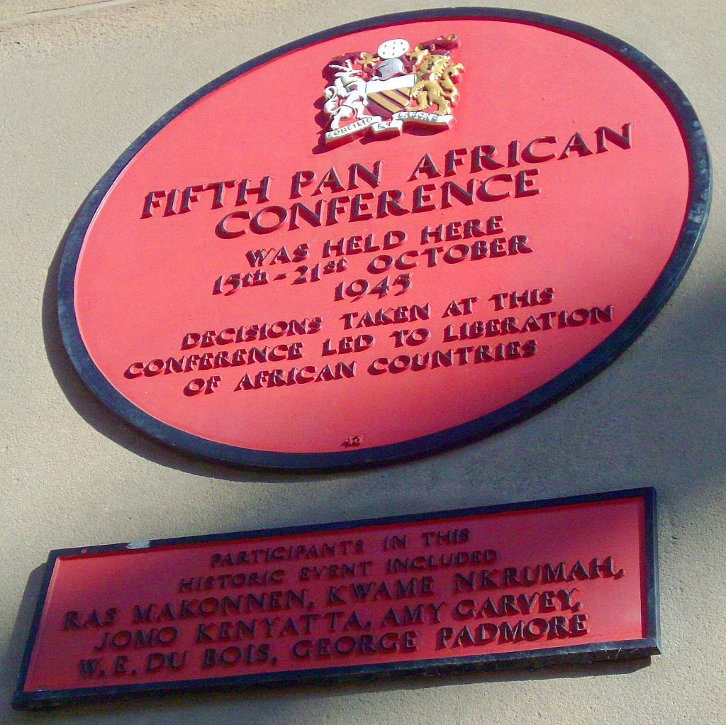 A red plaque on a concrete wall, with a navy blue outline. A lion insignia is visible at the top. It reads: "FIFTH PAN AFRICAN CONFERENCE. / WAS HELD HERE 15TH-21ST OCTOBER 1945 / DECISION TAKEN AT THIS CONFERENCE LED TO LIVERATION OF AFRICAN COUNTRIES". Below is a square plaque which reads: "PARTICIPANTS IN THIS HISTORIC EVENT INCLUDED / RAS MAKONNEN, KWAME NKRUMAH, JOMO KENYATTA, AMY CARVEY, W. E. DU BOIS, GEORGE PADMORE"
