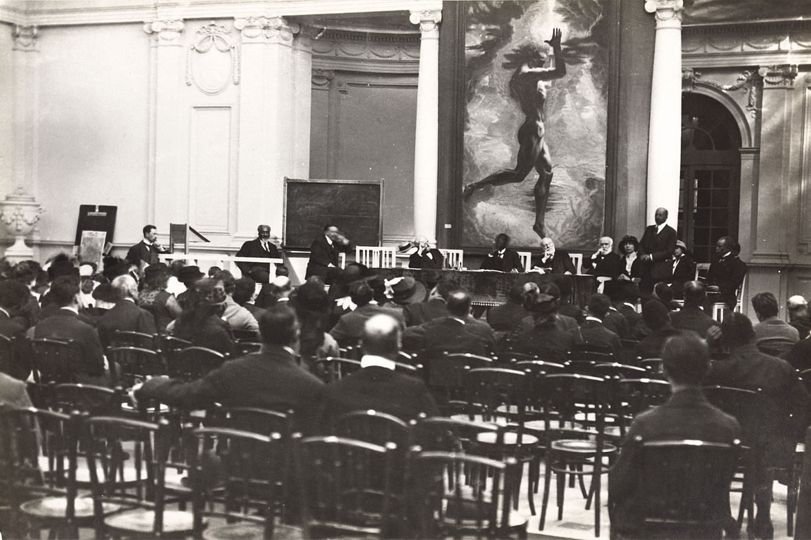 A black and white image of a crowd of people sat on rows of wooden chairs. The image is taken from behind, and they are looking towards a panel of speakers at the front. Above the speakers is a large painting of a muscled figure.