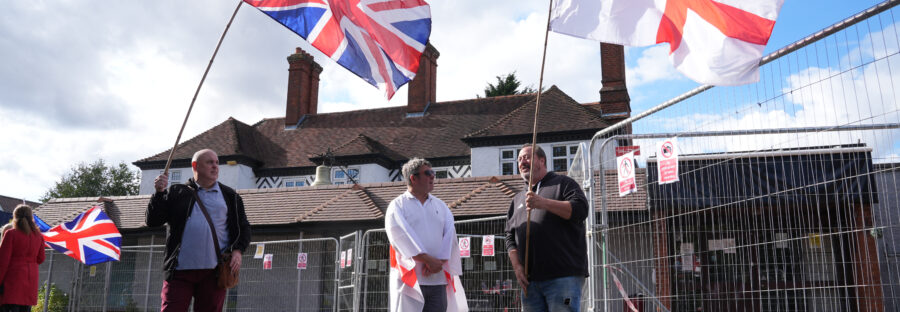 Three white men stand in front of a hotel on a sunny day. They are smiling and holding Union Jack and St George's flags.