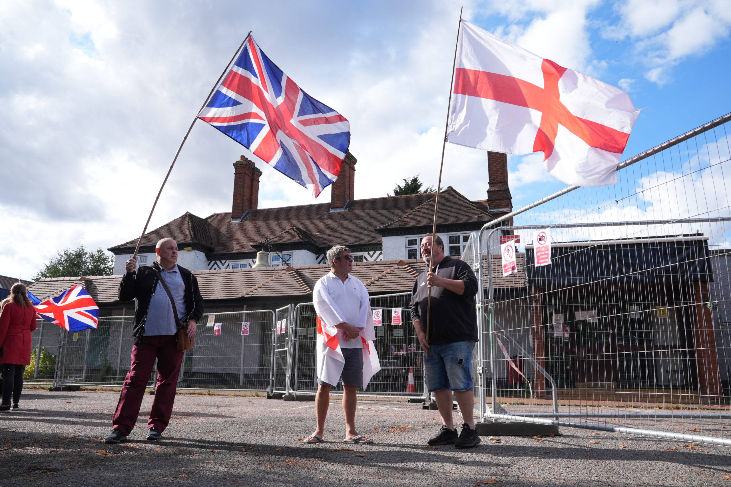 Three white men stand in front of a hotel on a sunny day. They are smiling and holding Union Jack and St George's flags.