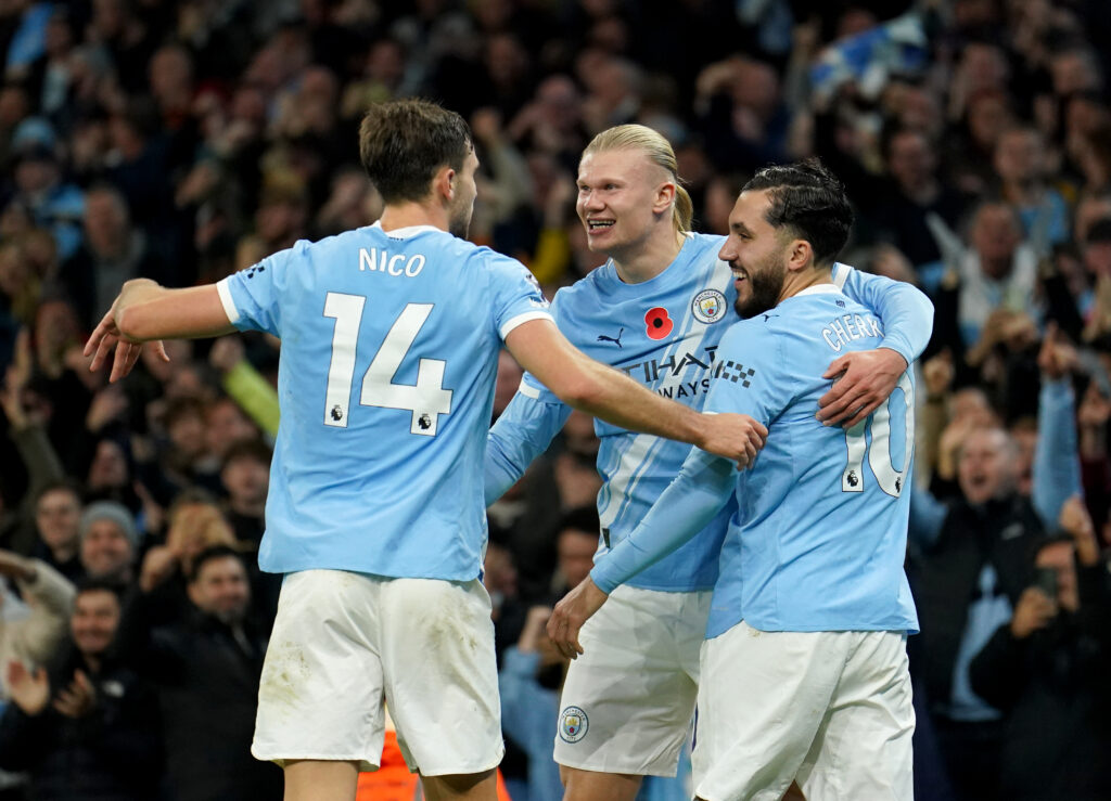 Three players in light blue Manchester City shirts celebrate in front of a crowd of cheering fans. 