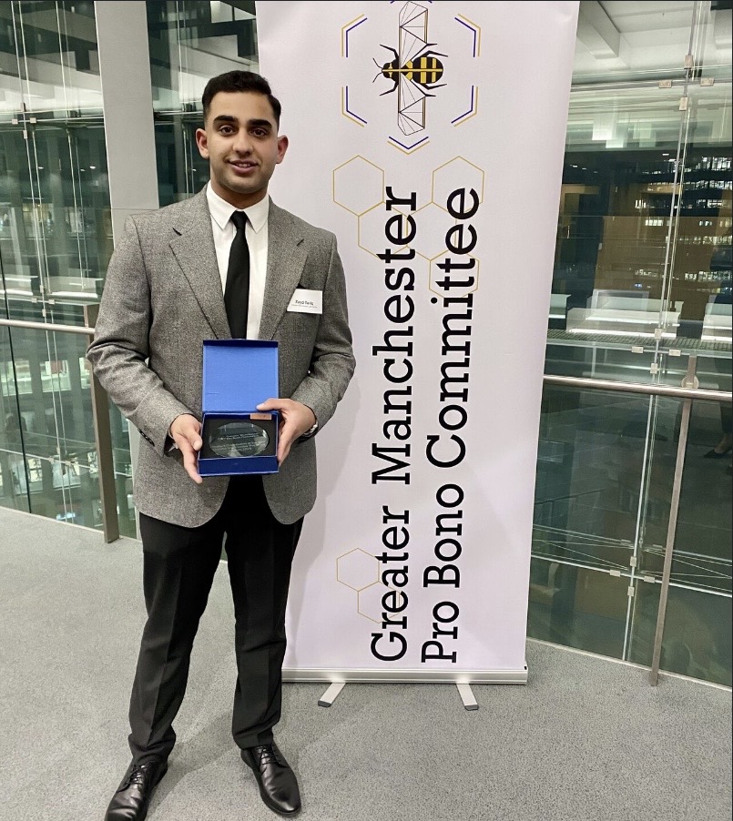 a man in a suit holding an award in front of a banner that says "Greater Manchester Pro Bono Committee"