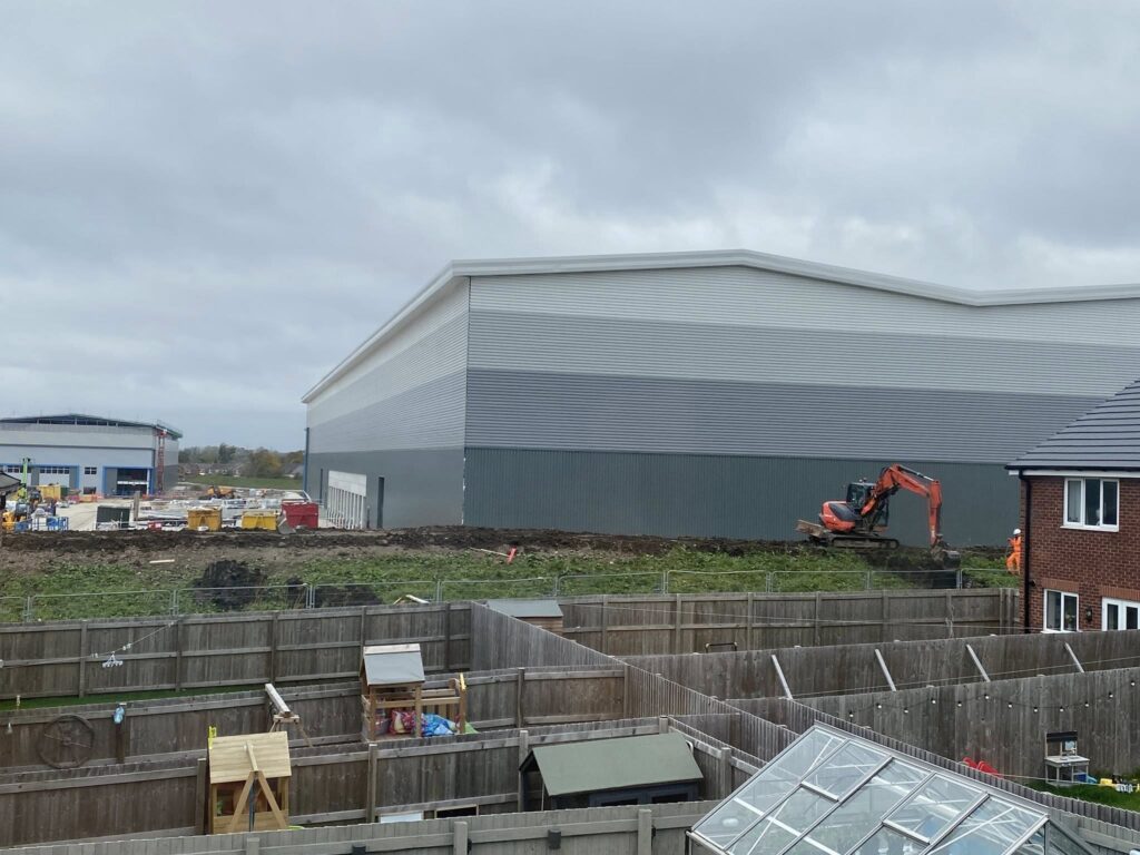 A large warehouse stands behind rows of residential gardens. Diggers and workmen are visible in the background.