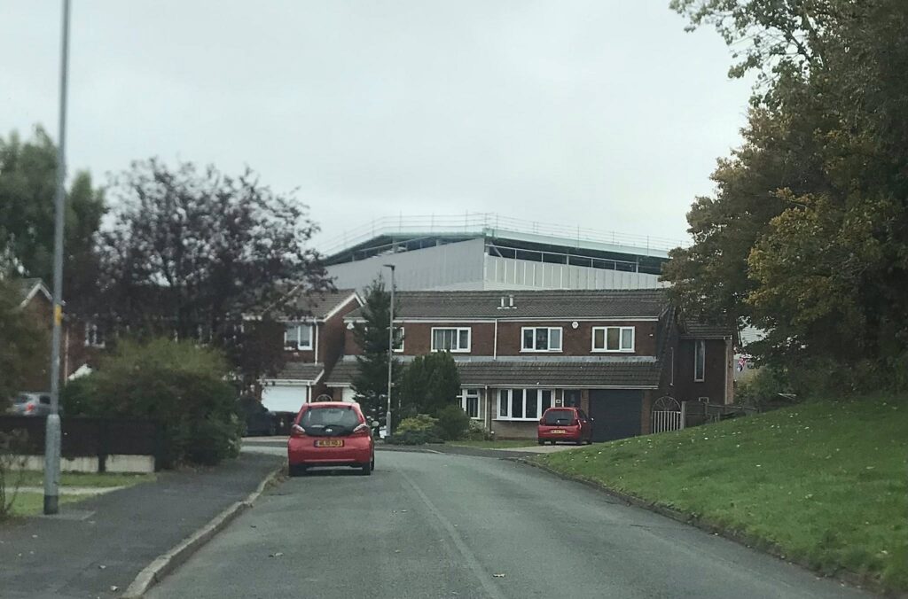 A picture of a residential street. There are trees and grass on either side of the road, and a row of housing in the background. A large, half-constructed warehouse is visible over the houses.